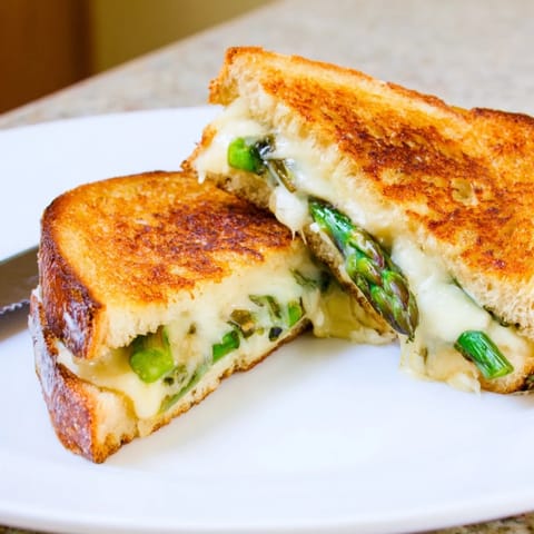 A close-up of the Asparagus & Brie Grilled Cheese reveals gooey Brie oozing between crusty bread and perfectly grilled, vibrant green asparagus spears.