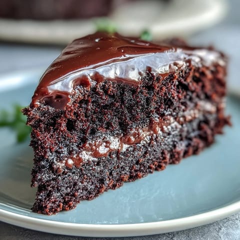 A slice of moist Easy Chocolate Fudge Cake on a white plate, showcasing a rich, fudgy texture and a glossy chocolate icing.